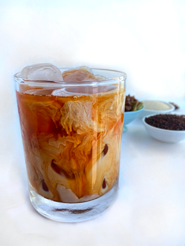 A cup of Shanti Chai & Co's Original Chai Blend served over ice, for a delicious glass of iced chai. Small bowls with tea and chai spices sit behind glass.