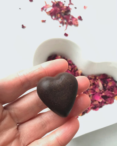 Shanti's hand holding a Vanilla and Rose Petal Chocolate, with a heart-shaped bowl full of red rose petals in the background.