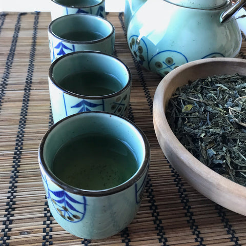 Cups of freshly brewed green tea sit beside a paired teapot and wooden bowl full of Sencha Green Tea leaves.