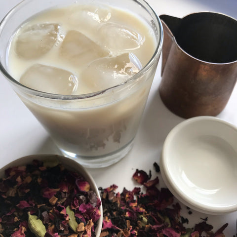A cup of Iced Chai, made with simple steep method. Shanti Chai & Co's Rose Petal Chai was used for this iced chai. A copper carafe for milk, a glass of iced chai, a white bowl of rose petal chai. 