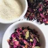 Raw sugar and Shanti Chai & Co's Rose Petal Chai sit in white bowls against a backdrop of rose petals and Assam tea. 