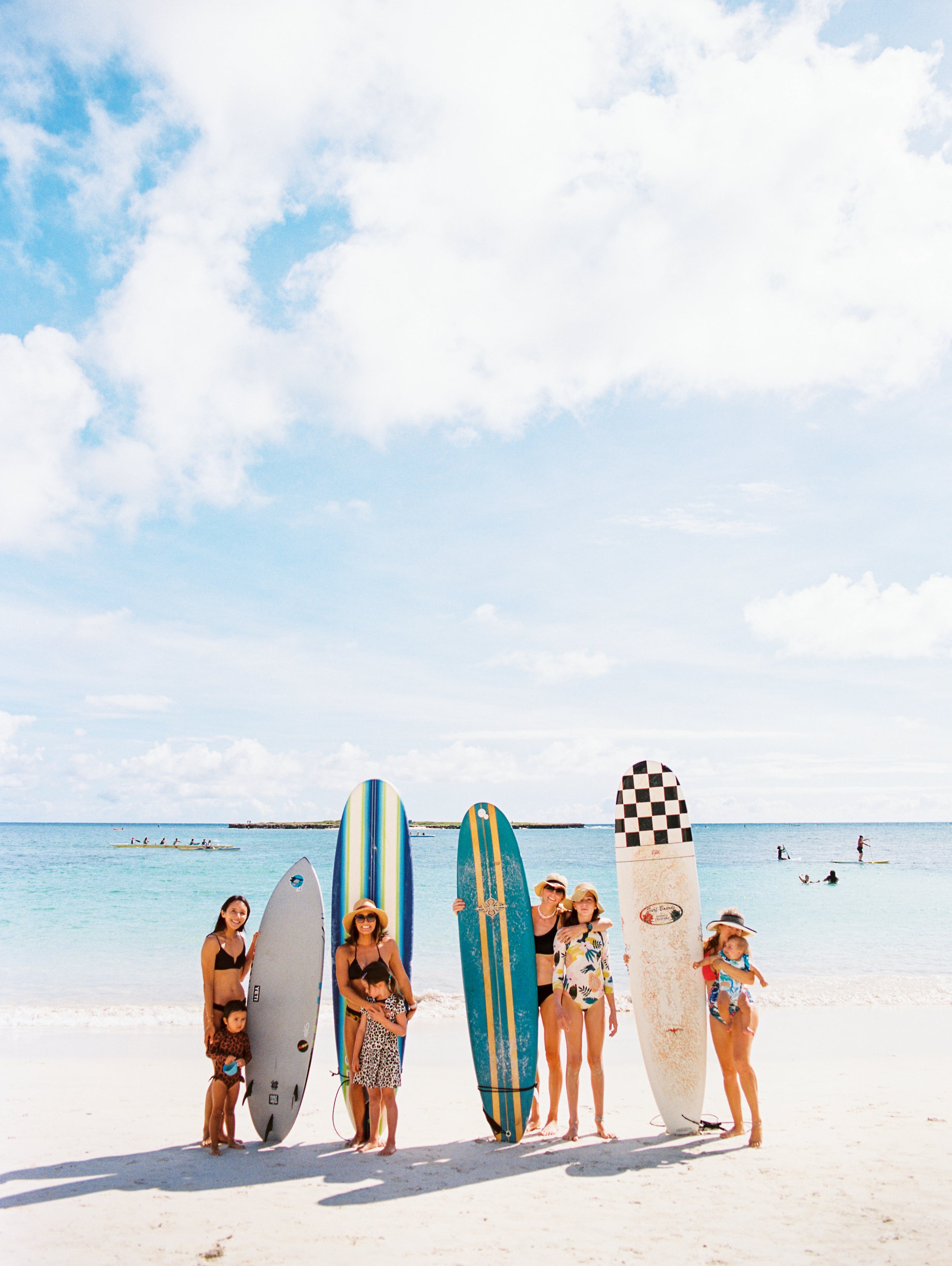 Group of women, members of Surfing Moms, posing on the beach with their kids and  surfboards.