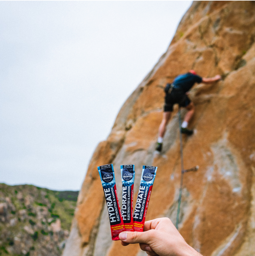 A hand holding electrolyte drink packets with a person rock climbing in the background.