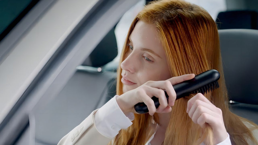 Red-haired woman in a white blouse using a black straightening brush in a car.