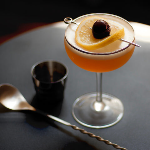 a Nightcap cocktail, which looks like a whiskey sour or an Amaretto sour and is garnished with a lemon slice and Luxardo cherry
