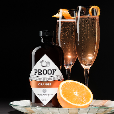 a bottle of Orange Proof Syrup on a silver tray next to half of an orange and two champagne cocktails