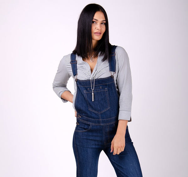 Indigo denim dungarees with grey top with hand on right hip.