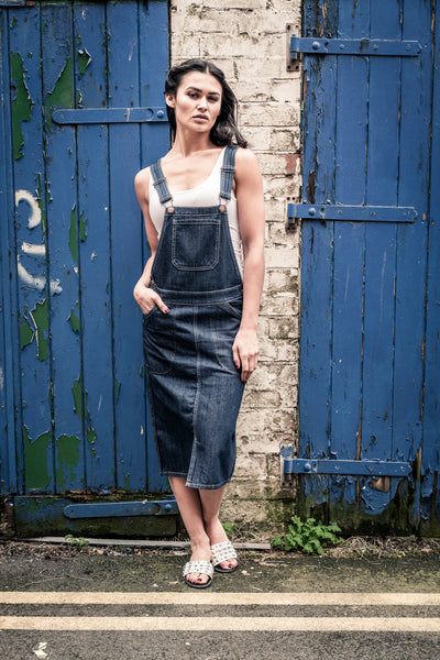 ‘Skylar’ style midi dark-wash dungaree dress in shabby urban alley.