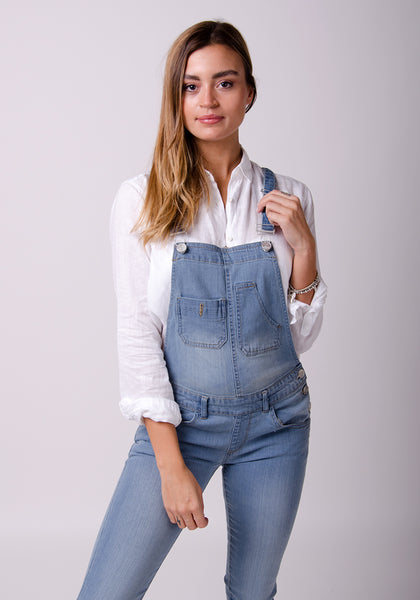 Pale wash skinny bib overalls for women paired with white loose-fit blouse while holding left strap.