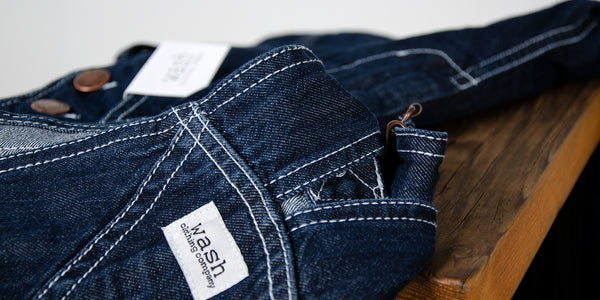 Close-up of dark denim dungarees with white stitching and Wash Clothing Company logo.