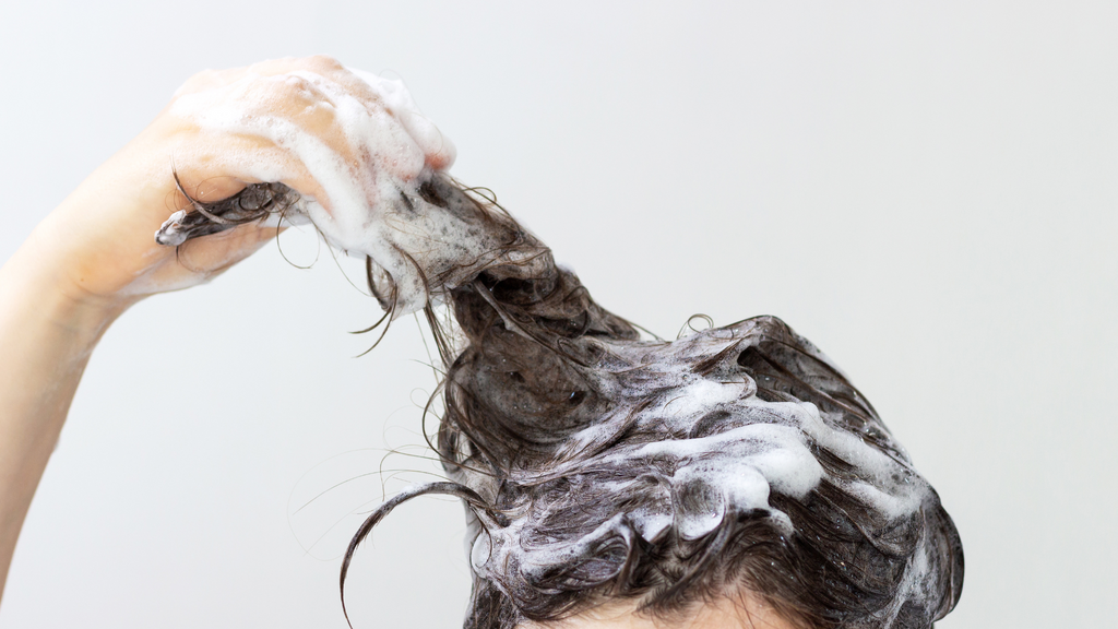 brunette woman washing her hair with shampoo foam in her hair holding a lock of hair with right hand