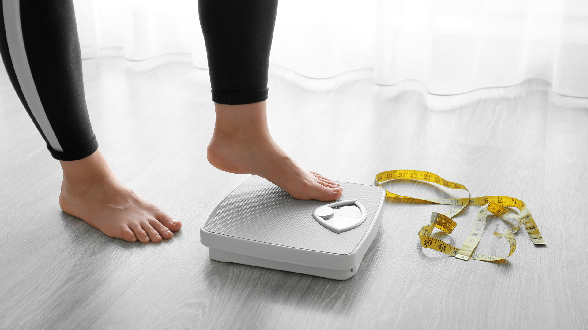 femme posant ses pieds sur une balance blanche, un mètre jaune posé à côté sur le sol