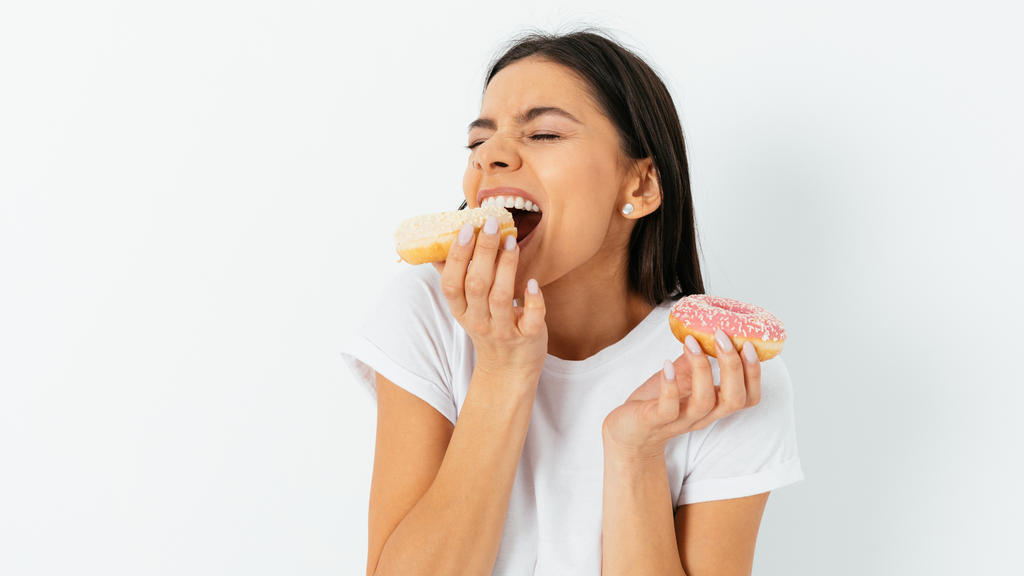 white skin brunette woman with a donuts in each hand. She bites into the one on the left with relish