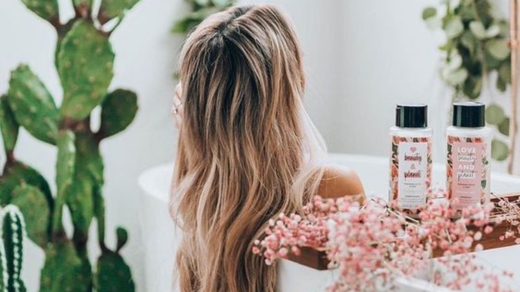 Blonde woman with long hair taking a bath, surrounded by hair care and plants (cacti)