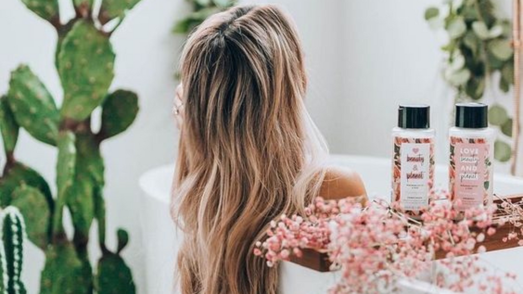 Femme blonde aux cheveux longs prenant un bain, entourée de soins capillaires et de plantes (cactus)