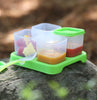 Green food cubes holding baby food on a rock outside with a Green Feeding Spoon placed beside.