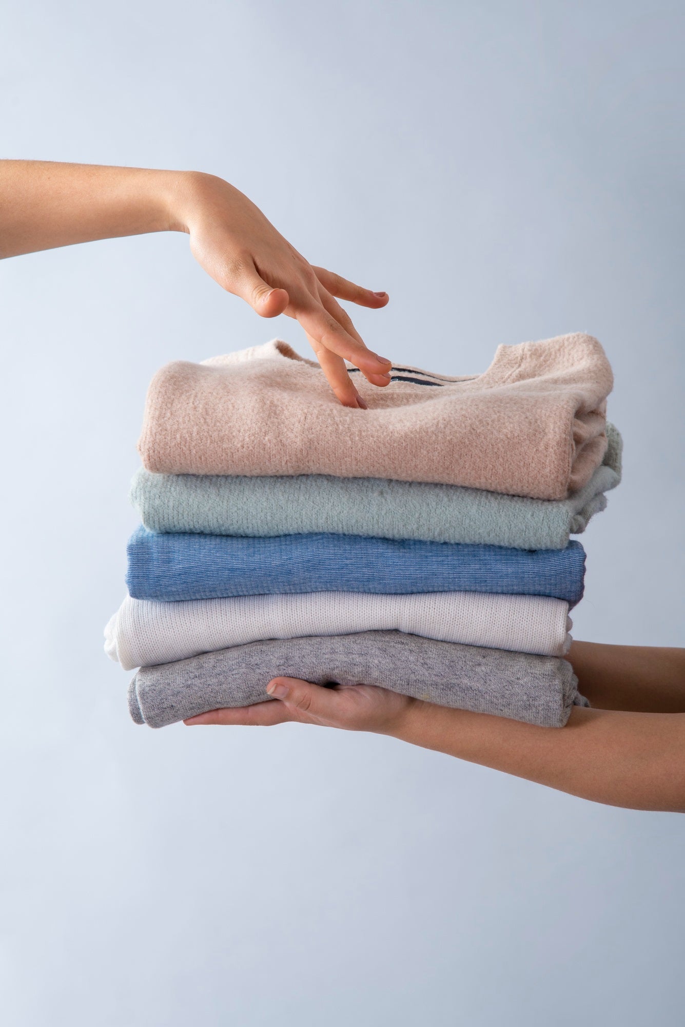A stack of neatly folded clothes in soft colors being held by one person while another hand reaches to pick a piece from the top of the pile.
