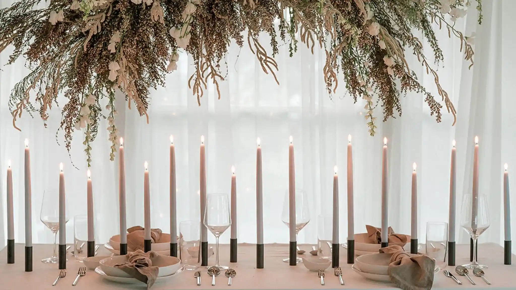 A row of lit candle sticks on a dinner table. There is a hanging plant decor piece hanging above and sheer white curtains in the background.