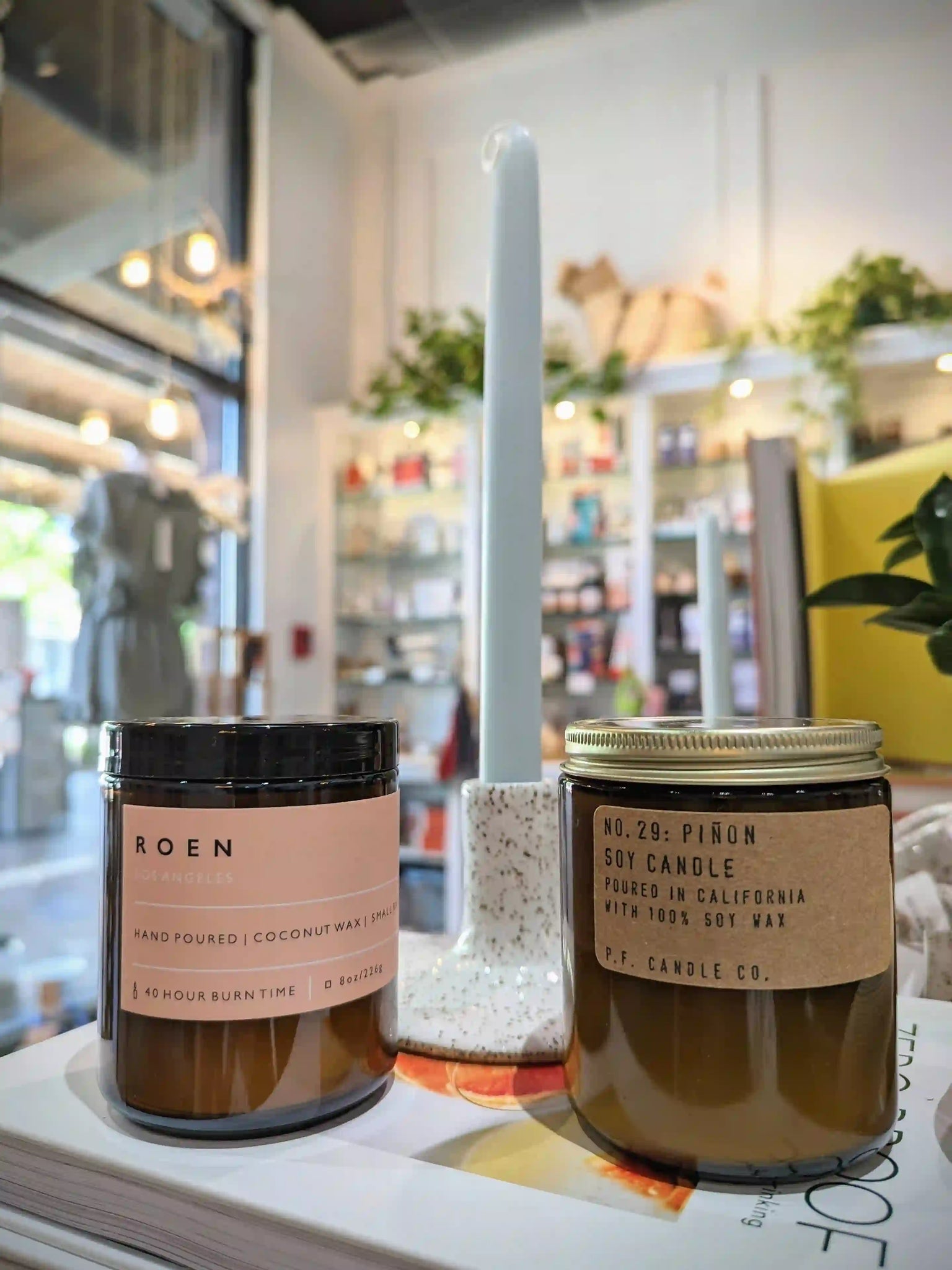 3 candles on top of a book with a store shelf in the background. The candle on the left is a brown jar with a pink label and black lid. The candle in the middle is a tall blue candle stick in a ceramic candlestick holder. The candle on the right is a brown jar with a brown label and a tin lid.