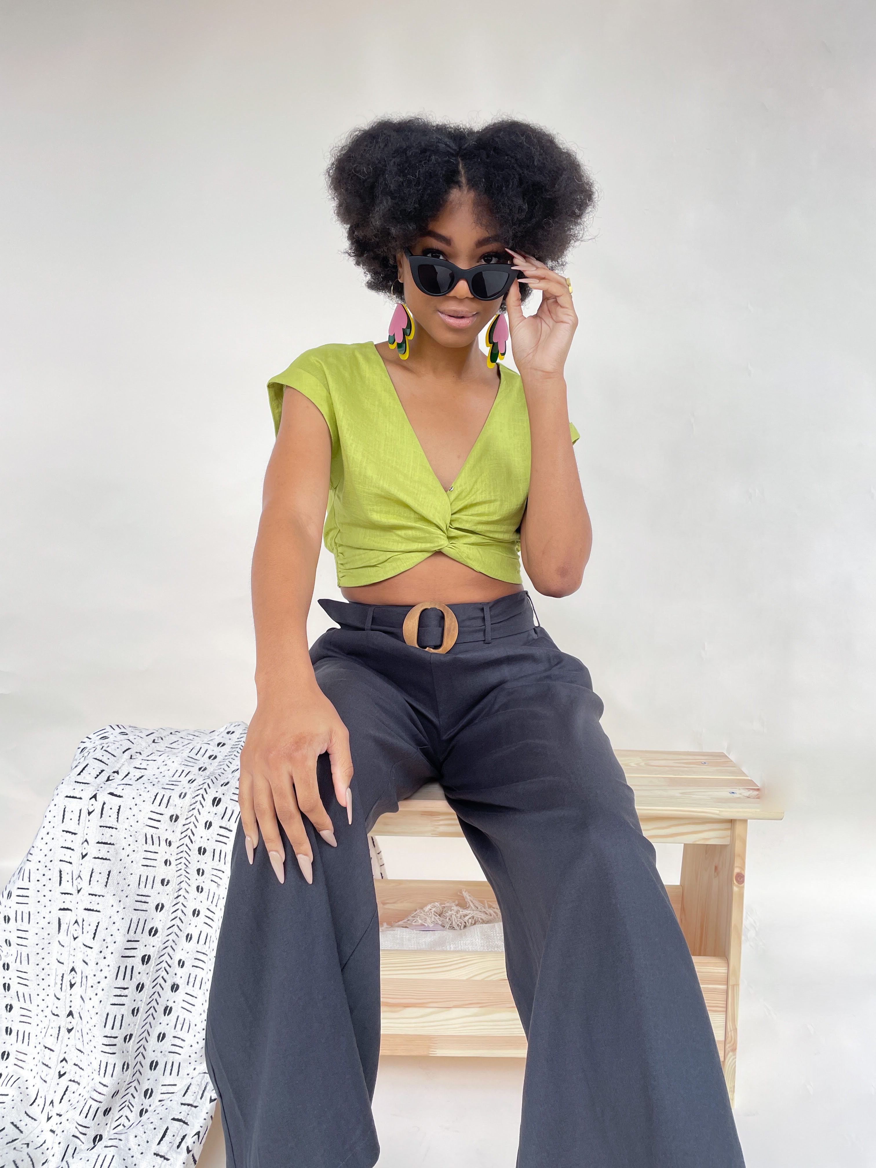 A woman with an afro wearing sunglasses, a green blouse, and black pants. She is sitting on a wooden bench with a blanket on it.