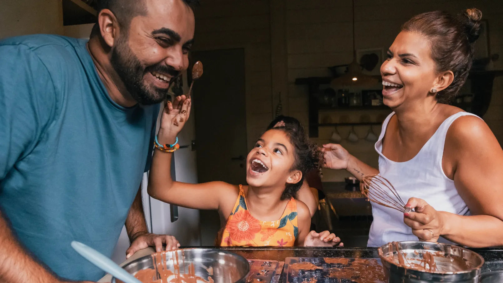 A family cooking and laughing. The dad is on the left, a daughter in the middle, the mom is on the right. The daughter is putting chocolate on the dad's face