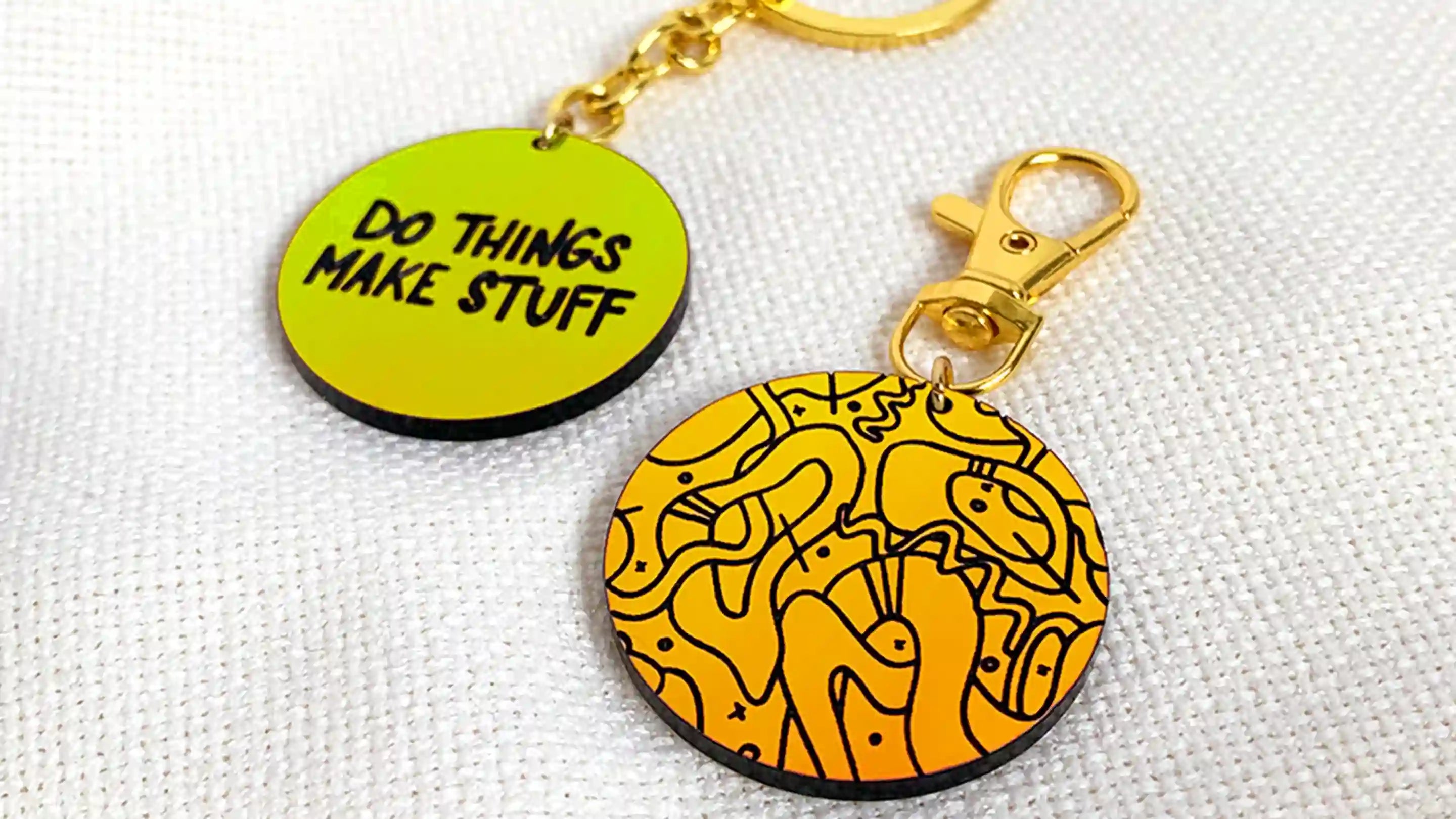 Two keychains placed next to each other on a piece of cloth. One is orange with an abstract design. The other is green and has the words "Do Things Make Stuff" on it.