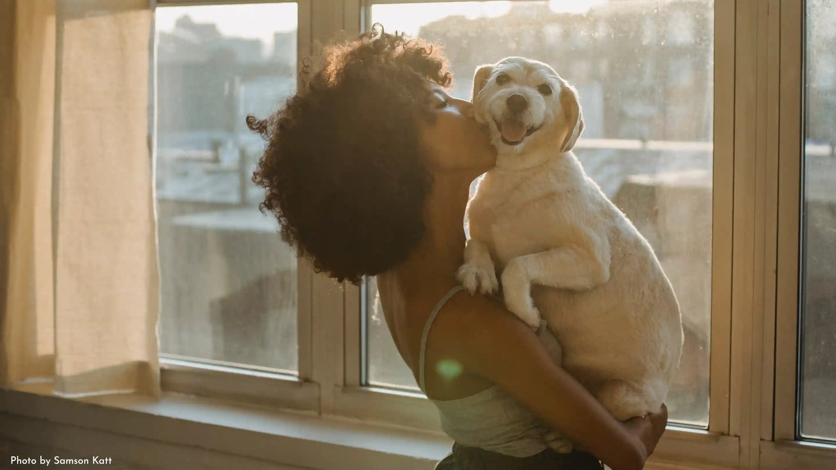 A black woman by a window kissing her dog on the cheek