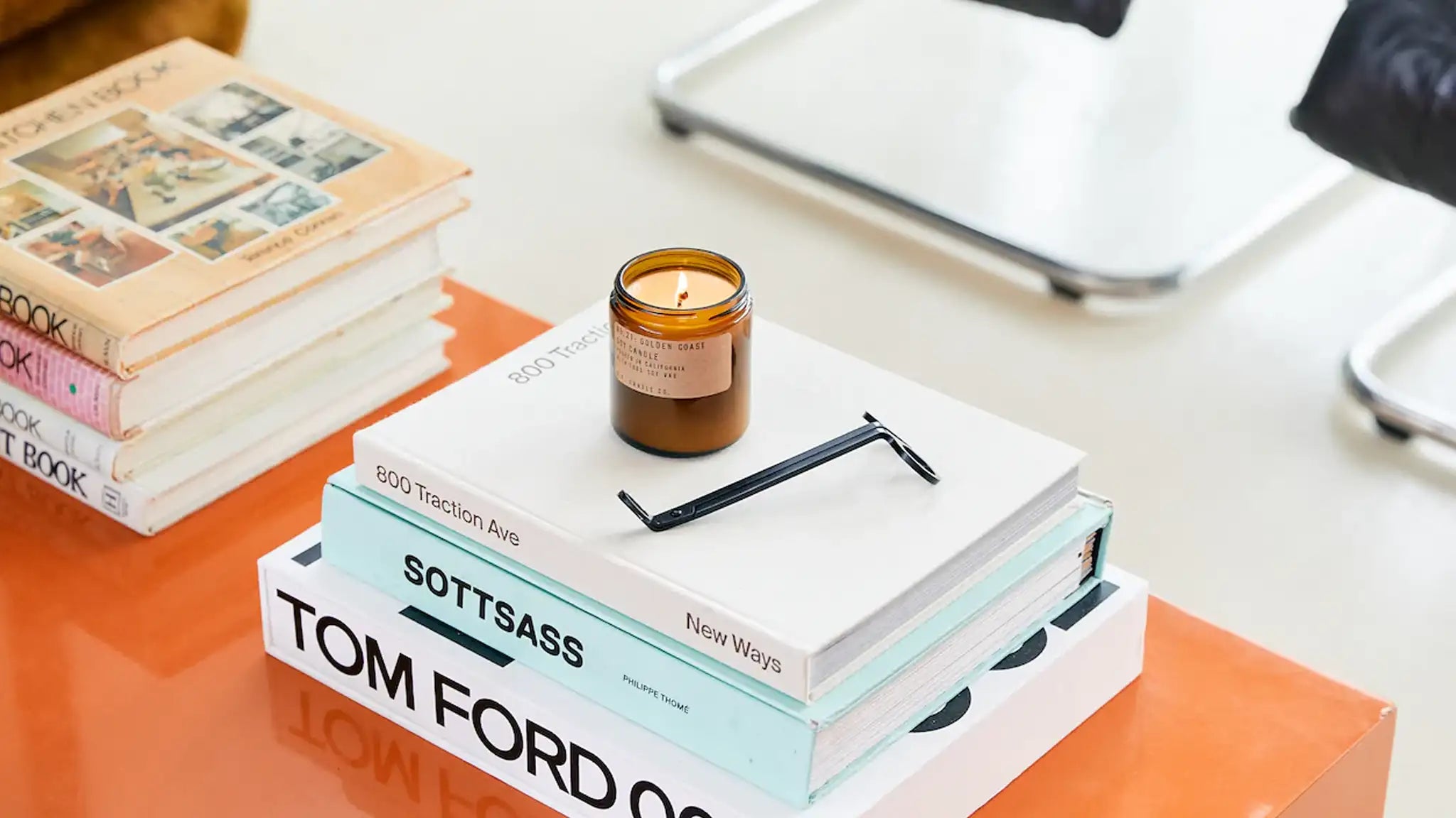 A lit candle jar next to a candle wick trimmer sitting on top of three books. The books are on top of an orange table next to more books. The angle of the photo was taken from above so the background is a white floor. 