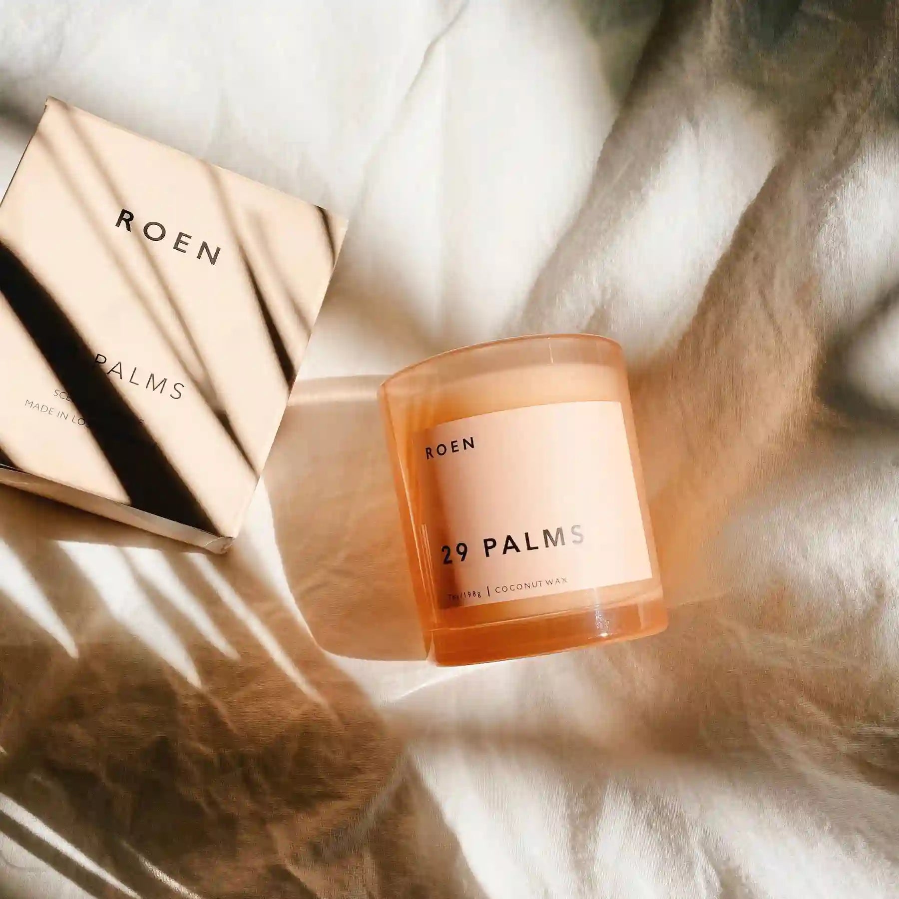 A candle in a pink glass jar with a pink packaging box resting on cloth