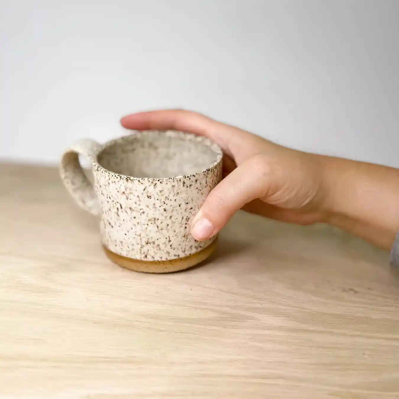 A hand holding a speckled ceramic mug. The mug is on a wooden table.