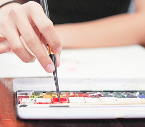 hand dipping paintbrush into watercolor paint pan