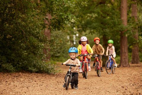 a two year old child riding a bike through a forest with other kids behind him