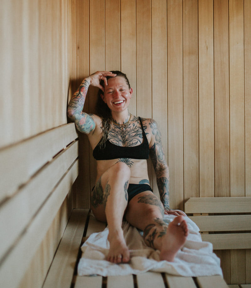 Woman sitting on a sauna bench laughing while she uses the sauna for workout recovery