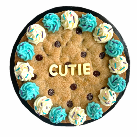 Image shows an isolated photo of our signature cookie cake. blue and white swirly icing surrounds it and the word "CUTIE" is in the middle, written with white chocolate blocks