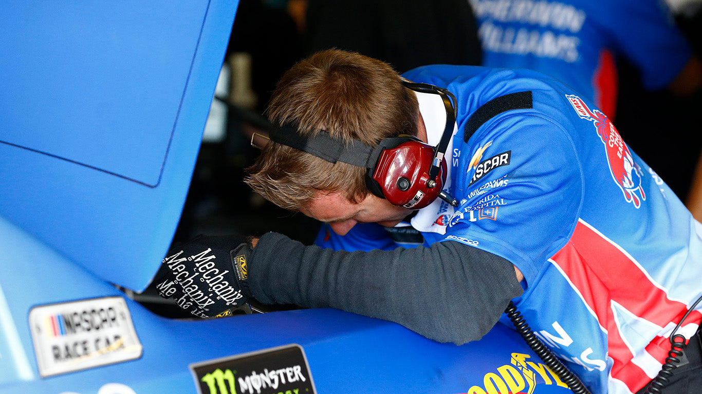 The image captures a moment of intense concentration as a NASCAR technician, clad in a vibrant blue team uniform and wearing Mechanix Wear The Original Red Work Gloves, leans into a race car's internals. The prominent display of the gloves with their distinctive branding highlights their use in high-stakes, precision-demanding environments. This real-world application showcases the gloves' suitability for tasks requiring both dexterity and protection. The alt text and description are crafted to enhance search engine visibility with relevant keywords while ensuring accessibility for users with visual impairments, describing the scene in clear, engaging language.