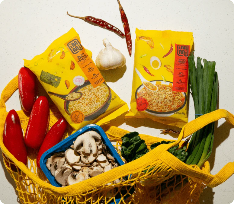 Ramen noodle packages with fresh vegetables and spices in a yellow net bag.