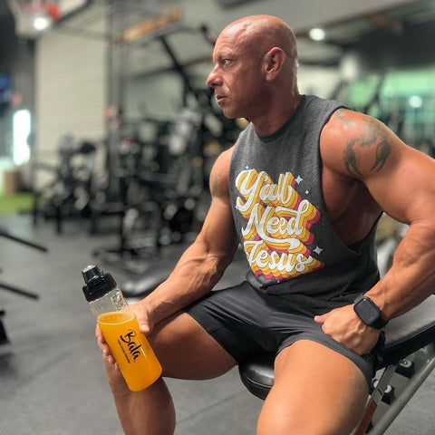 A muscular man in a gym sits on a bench holding a bottle of Bala Total Body Wellness Drink Mix by Bala Enzyme, an orange liquid packed with electrolytes. He wears a sleeveless shirt with the text "Y'all Need Jesus," black shorts, and a smartwatch. Gym equipment is visible in the background.