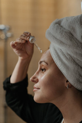 A woman in front of a mirror with a towel on her hair applying an oil on her skin