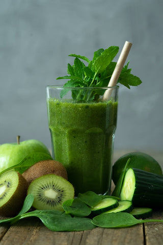 A glass of green smoothie with few pieces of Kiwi and spinach leaves around it