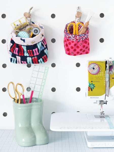 Two fabric baskets made from fabric scraps hanging from a pegboard above scissors and crafting supplies on a table