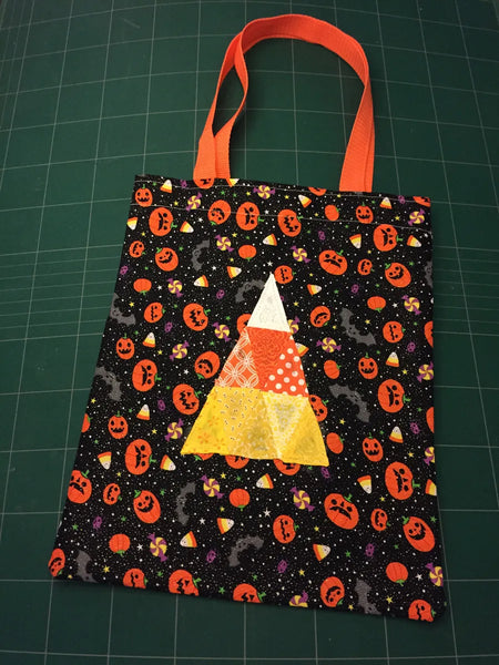 An English Paper Pieced candy corn design composed of triangles in orange, yellow and white is stitched to a bag made of orange and black Halloween fabric