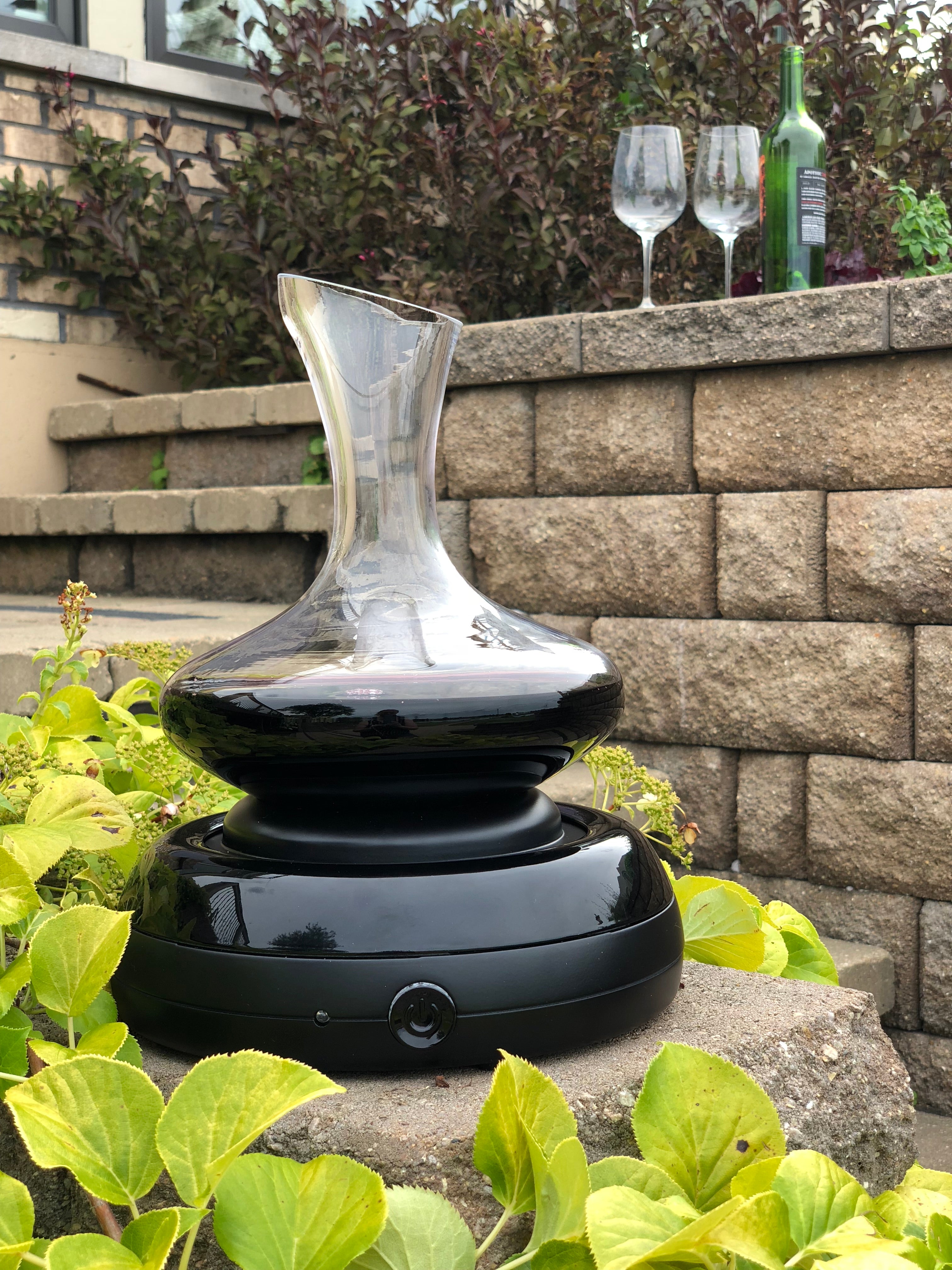 Aerisi Wine Aerator and Decanter with red wine sitting on patio sone stairs surrounded by lush green vines with wine bottle and 2 wine glasses in background