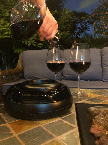 Aerisi wine aerating unit on patio table near fire pit with blue outdoor couch in background, hand holding decanter and pouring red wine into 2 wine glasses