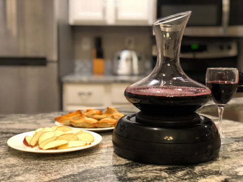 Aerisi Wine Aerator with red wine in decanter and a glass of red wine standing to the right of the Aerisi unit sitting on a white and black granite kitchen counter next to a plate of sliced apples.top 