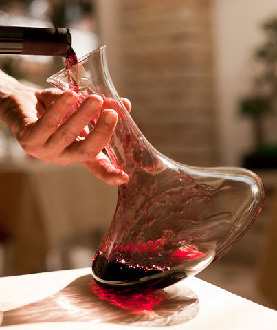 Hand tilting wine decanter on wooden table pouring a bottle of red wine flowing along sides of glass decanter to aerate