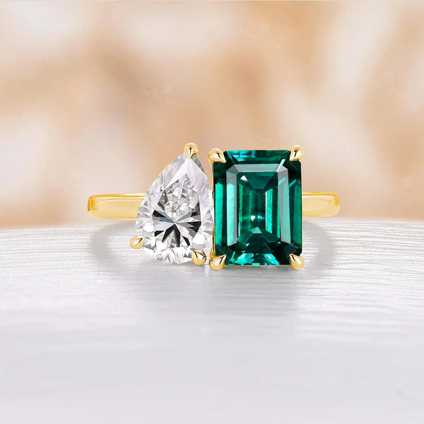 A gold ring featuring a pear-shaped moissanite gemstone and an emerald-cut green gemstone, elegantly displayed on a light surface with a soft, blurred background.