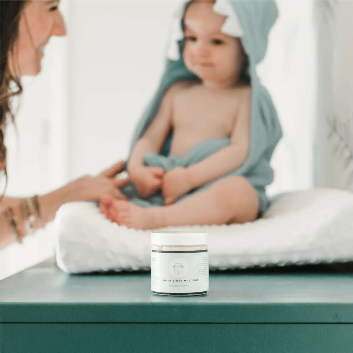 A baby wrapped in a towel sits on a changing table with a lotion jar in the foreground.