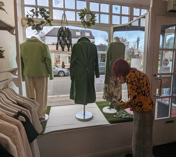 A woman is measuring out decorative plants behind a shop window. The window is full of mannequins dressed in green clothing, artificial grass and plants.