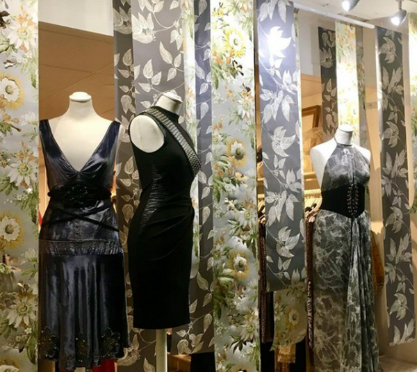 A shop window displaying evening dresses on mannequins next to strips of floral printed wallpaper.