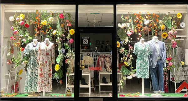 A double fronted shop window displaying floral dresses amongst brightly coloured decorative flowers.